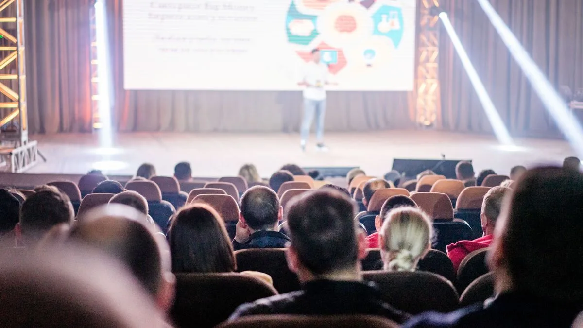 Audience attentively watching a presentation in a conference setting, with stage lights and speaker visible.